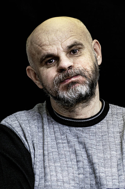 Porträt eines glatzköpfigen Mannes mit grauem Bart und geschwollenen Wangen auf dunklem Hintergrund - Foto, Bild