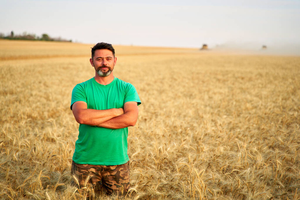 Happy farmer proudly standing in wheat field with arms crossed on chest. Agronomist wearing corporate uniform, looking at camera on farmland. Rich harvest of cultivated cereal crop. Harvesting season. - Fotoğraf, Görsel