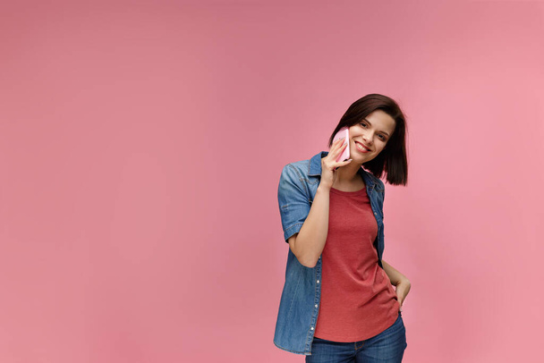Young people working with mobile devices. Portrait of cute happy brunette woman wearing t-shirt and jeans shirt talking on mobile phone and smiling isolated over pink background. Copy space - Fotoğraf, Görsel