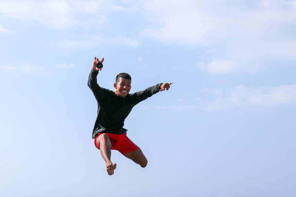 Homem pulando feliz na praia com um céu azul no fundo. Jovens saltos indonésios alegres. Um adolescente de bom humor salta para o ar. Fundo com céu azul. Dia tropical ensolarado
. - Foto, Imagem