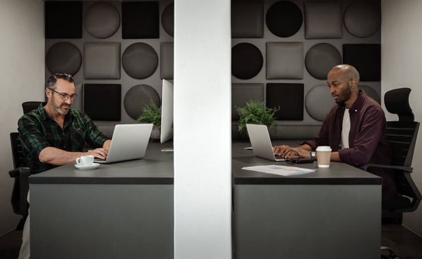 Two diverse businessman working on laptops in separate cubicles while physical distancing in the workplace - Photo, Image