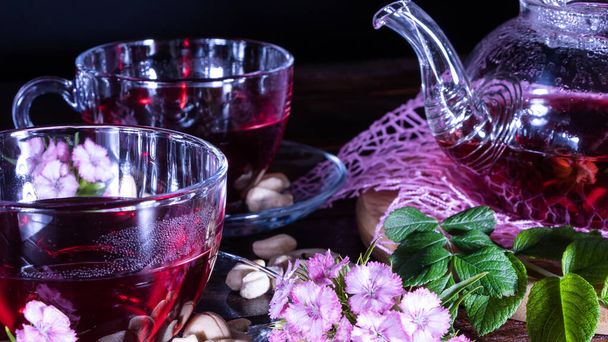 A mug of red tea and a teapot in white hibiscus flowers and green leaves of medicinal tea on a wooden stand.Zen tea ceremony. Photo of red herbal Indian healing tea.Elegant mugs with a relaxing drink - Photo, Image