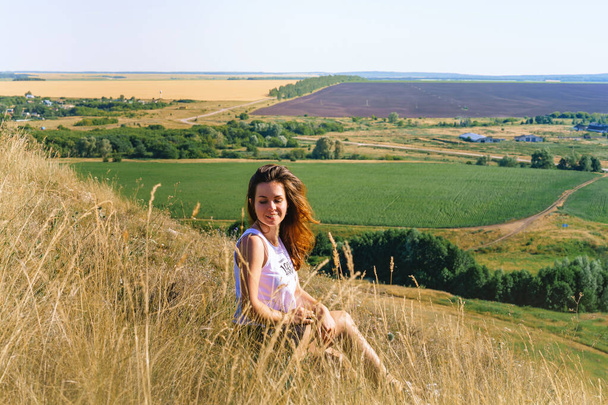 Vrouw toerist met lang haar zit op het gras in velden die eruit zien als Toscane met uitzicht op eindeloze weiden en bergen - Foto, afbeelding