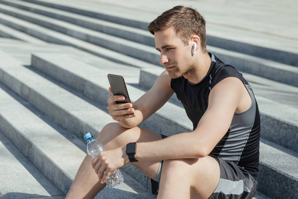 Athlete with wireless headphones and fitness tracker holds bottle of water and smartphone in hands, sits on stairs - Foto, imagen