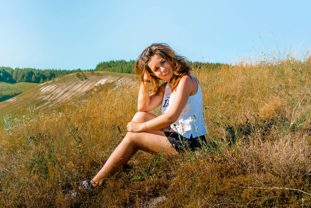 Uzun saçlı bir kadın turist, uçsuz bucaksız çayır ve dağ manzaralı Toskana 'ya benzeyen tarlalarda çimenlerde oturur. - Fotoğraf, Görsel