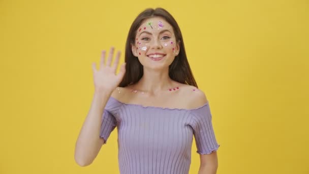 A friendly young girl with fashion makeup and stickers on the face is waving to the camera doing hello gesture isolated over a yellow background - 映像、動画