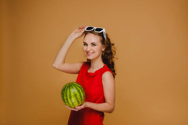 Photo of a cute girl in a red dress smiling with a watermelon and melon in her hands isolated on a bright red background - Фото, изображение