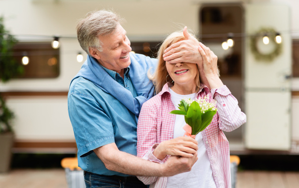 Unexpected surprise. Senior man closing his wifes eyes and giving her flowers at campground - 写真・画像