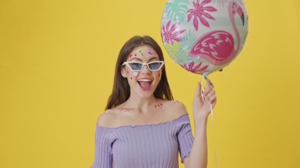 A cheerful young girl with fashion makeup and stickers on the face releases up the balloon standing isolated over a yellow background - Séquence, vidéo