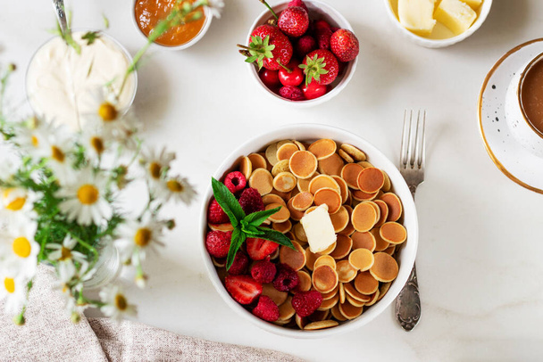 Mini pannenkoeken cornflakes met bessen en muntblaadjes op witte achtergrond. Zure room en jam. Een hamomile. Trendy Food breakfast Concept. Bovenaanzicht. Kopieer ruimte voor tekst. - Foto, afbeelding