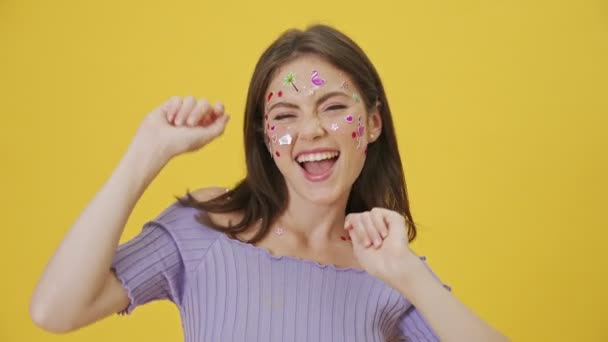 A cheerful young girl with fashion makeup and stickers on the face is dancing standing isolated over a yellow background - Filmati, video