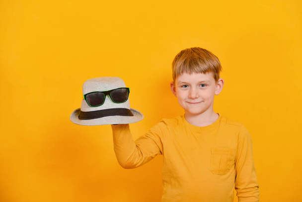 Un niño con una camiseta naranja sostiene un sombrero gris con gafas en la mano derecha
. - Foto, imagen