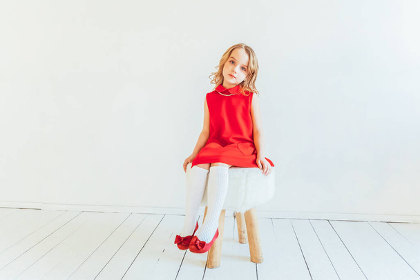 Γλυκό κοριτσάκι με κόκκινο φόρεμα κάθεται στην καρέκλα ενάντια στο λευκό τοίχο στο σπίτι, χαλαρώνοντας σε λευκό φωτεινό σαλόνι σε εσωτερικούς χώρους. Η νεολαία της παιδικής ηλικίας χαλαρώνει έννοια - Φωτογραφία, εικόνα