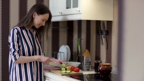 Mulher feliz cortando pepino na cozinha. Vídeo. Vista lateral de uma jovem bonita com cabelo escuro longo e camisa listrada cozinhar prato vegetariano, conceito de dieta saudável
 - Filmagem, Vídeo