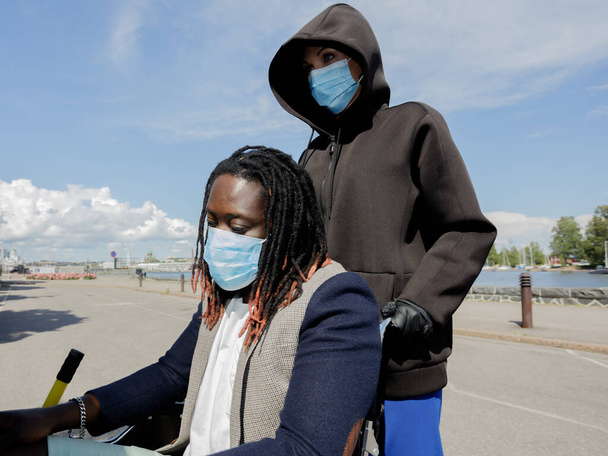 Las mujeres europeas y los hombres africanos llevan una máscara médica para protegerse del brote de coronavirus. Primer plano. Un hombre en silla de ruedas. Día soleado de verano. Camina por el terraplén
. - Foto, imagen