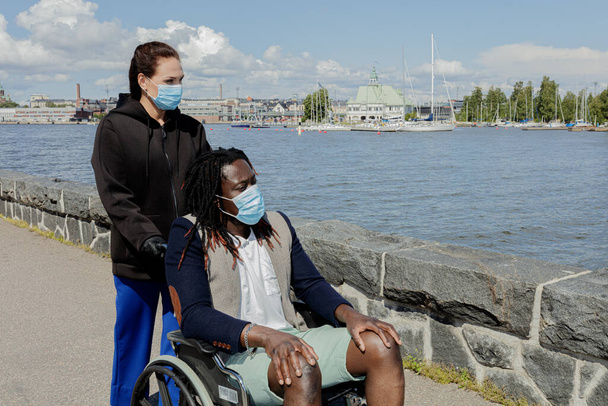 Les femmes et les hommes africains européens portent un masque médical pour se protéger de l'épidémie de coronavirus. Gros plan. Un homme en fauteuil roulant. Journée ensoleillée d'été. Promenade le long du remblai. - Photo, image