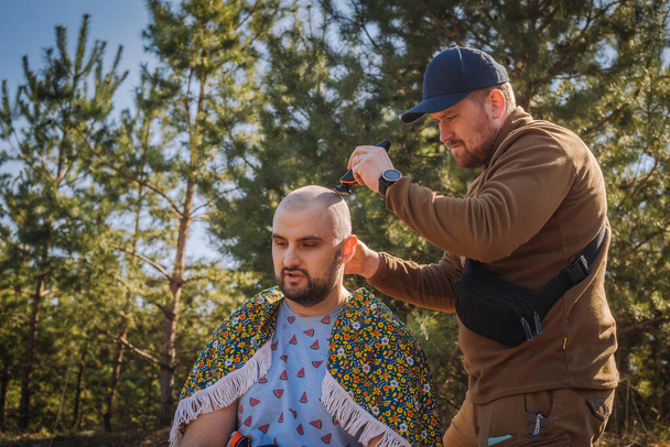 Tough red bearded guy shaving bold the other man outdoors in the pine forest - Photo, Image
