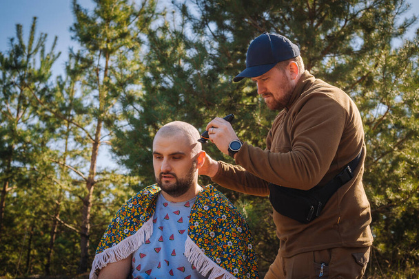 Kemény vörös szakállú fickó borotválja a másik férfit a fenyőerdőben. - Fotó, kép