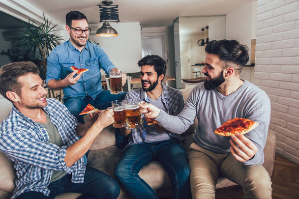 Четверо друзей-мужчин пьют пиво и едят пиццу дома. - Фото, изображение
