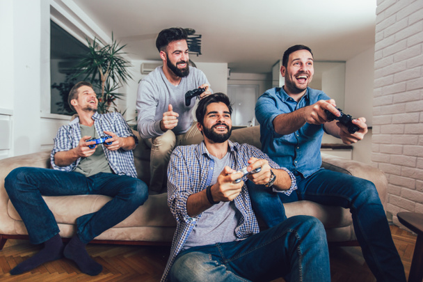 Hymyilevät miespuoliset ystävät pelaavat videopelejä kotona ja pitävät hauskaa. - Valokuva, kuva