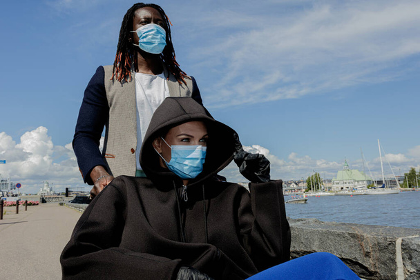 Las mujeres europeas y los hombres africanos llevan una máscara médica para protegerse del brote de coronavirus. Mujer en silla de ruedas. Día soleado de verano. Camina por el terraplén
. - Foto, Imagen