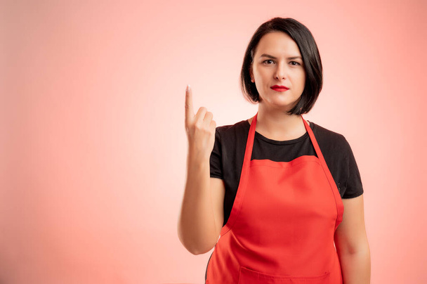 Kobieta zatrudniona w supermarkecie w czerwonym fartuchu i czarnym t-shircie, licząc jedną z palcem odizolowanym na czerwonym tle - Zdjęcie, obraz