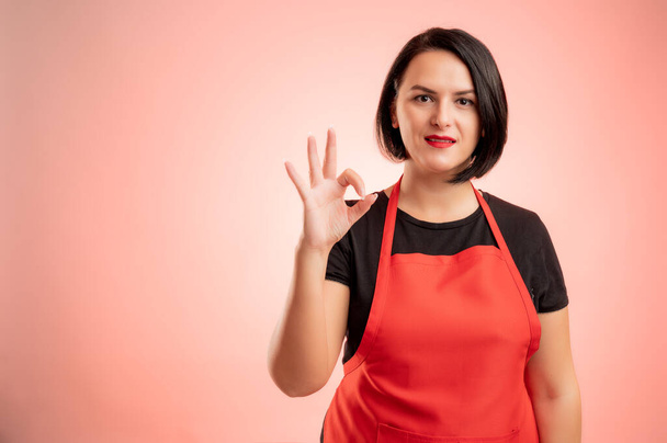 Woman employed at supermarket with red apron and black t-shirt, showing ok isolated on red background - Photo, Image
