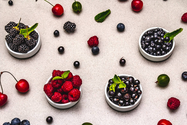 Ποικιλία μούρα σε κύπελλα: bilberry, blueberry, φραγκοστάφυλο, βατόμουρο, κεράσι και βατόμουρο. Πέτρα φόντο τσιμέντο - Φωτογραφία, εικόνα