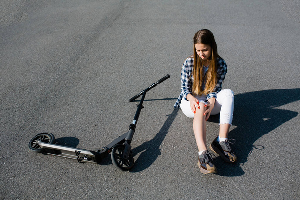 A girl sits on the asphalt next to an abandoned scooter and checks her knee for a bruise due to a fall from a vehicle onto the asphalt - Photo, image