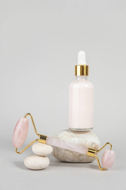 Crystal rose quartz facial roller and anti-aging collagen, serum in glass bottle on stones, grey background. Facial massage for natural lifting, Beauty concept Front view. - Valokuva, kuva