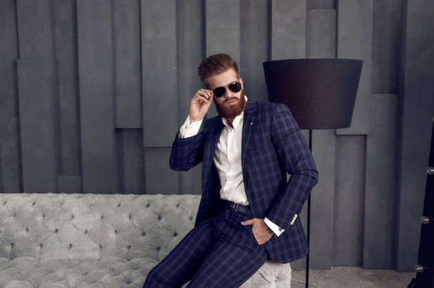 Handsome stylish man in blue suit in a cage and eyewear at home sitting on sofa - Valokuva, kuva