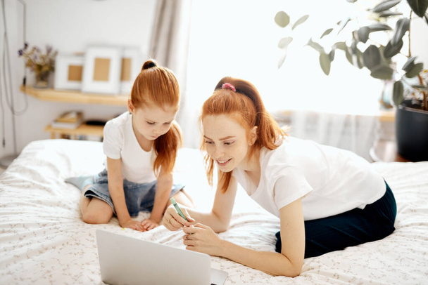 Red-haired Mom and her daughter lying on bed at home while looking at laptop - Foto, Imagem