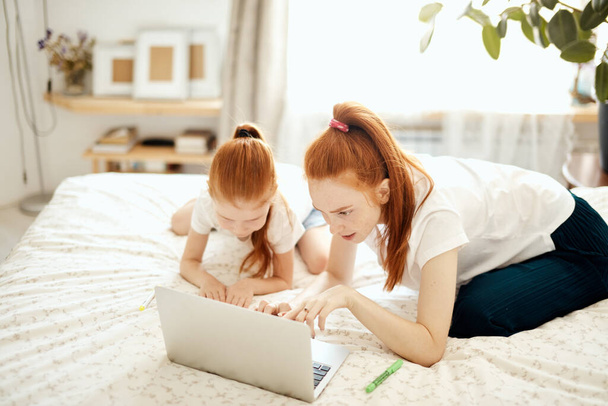 Red-haired Mom and her daughter lying on bed at home while looking at laptop - 写真・画像