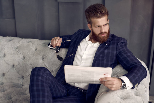 Przystojny stylowy mężczyzna w niebieskim garniturze w klatce w domu, siedzący na kanapie i czytający świeżą gazetę. - Zdjęcie, obraz
