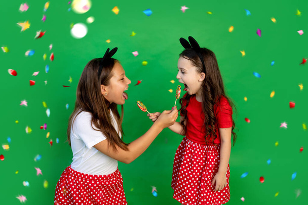 Cute little girls in red Mickey mouse outfits on a green isolated background with streamers eating large lollipops and showing their tongues. Space for text.holiday concept - Photo, Image