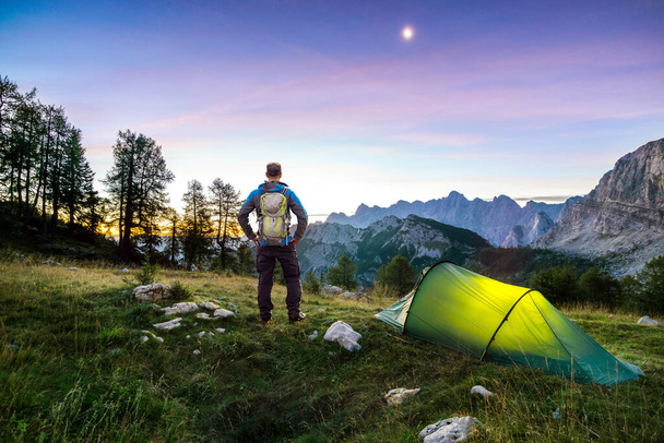 Hiker with Backpack standing on Mountain and Tent glowing under a moon night sky at sunset or sunrise twilight hour. Alps, Sleme, Triglav National Park, Slovenia. - Photo, Image