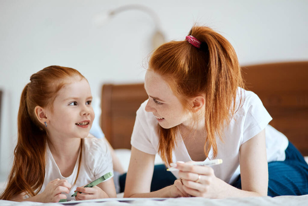 Red haired Mother lies on the bed with her preschool daughter and draws. - Valokuva, kuva