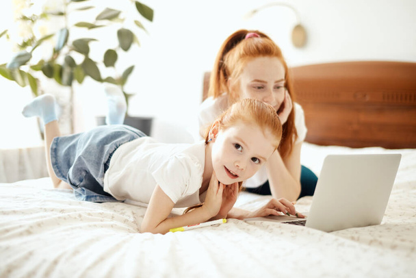 Red-haired Mom and her daughter lying on bed at home while looking at laptop - Fotó, kép