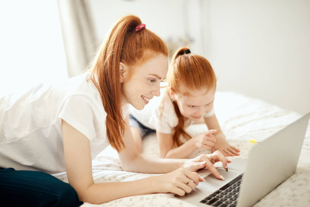 Red-haired Mom and her daughter lying on bed at home while looking at laptop - Φωτογραφία, εικόνα