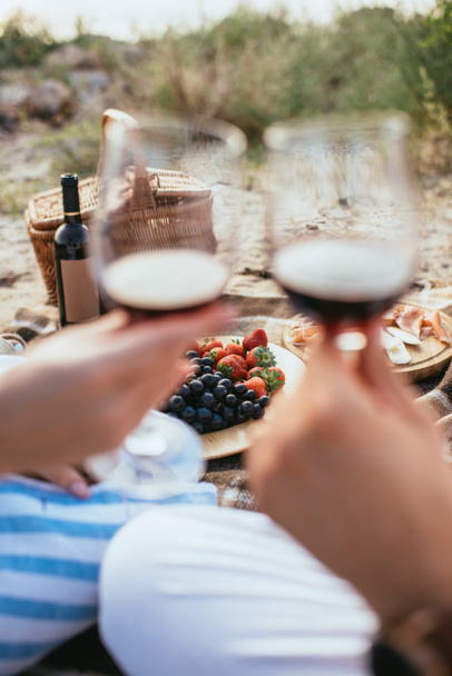 selective focus of plate with fresh fruits near bottle and couple clinking glasses with red wine  - Foto, immagini