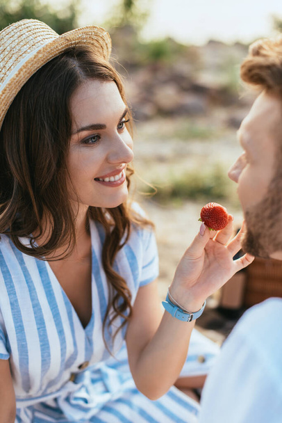 Selektiver Fokus der glücklichen Frau mit Strohhut, die Freund mit Erdbeere füttert  - Foto, Bild