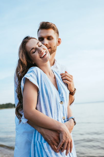 brodaty mężczyzna uśmiecha się i przytula wesoły dziewczyna w pobliżu jeziora - Zdjęcie, obraz