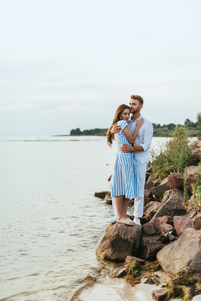 handsome man hugging beautiful woman in dress standing on rocks near river  - Photo, Image