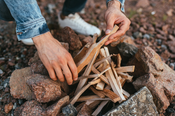 cropped view of young man touching sticks while making bonfire near stones  - Fotó, kép