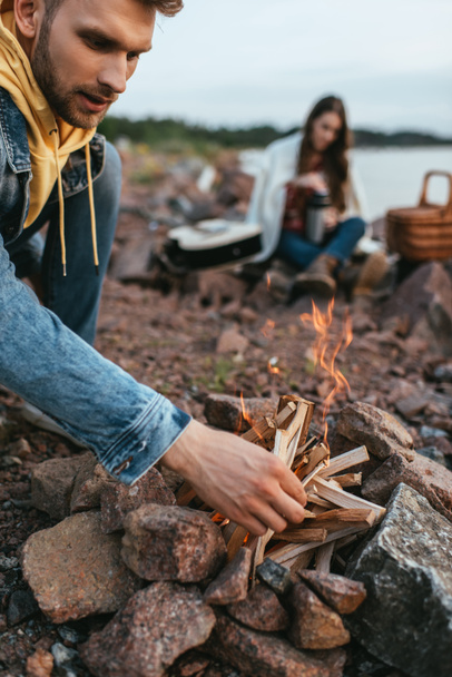 selective focus of handsome man touching sticks while making bonfire near woman  - Fotografie, Obrázek