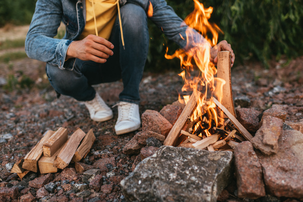 Ausgeschnittene Ansicht eines Mannes, der sitzt und Holz in brennendes Lagerfeuer legt  - Foto, Bild