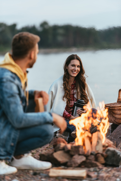 selective focus of happy woman holding thermos and looking at boyfriend near bonfire   - Photo, Image
