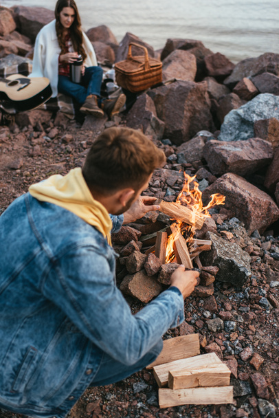 selective focus of man sitting near logs, burning bonfire and girlfriend  - Photo, Image