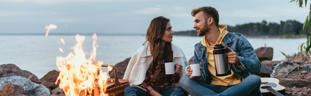 panoramic shot of couple holding cups and sitting near bonfire and lake - Foto, imagen