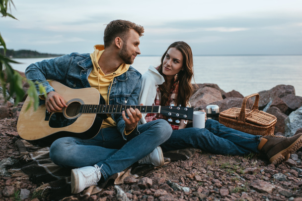 beautiful woman looking at handsome boyfriend playing acoustic guitar outside  - Fotoğraf, Görsel